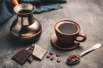 Black coffee drink in a clay cup, turkish jezve coffee pot, chocolate pieces and coffee beans in a spoon on dark background