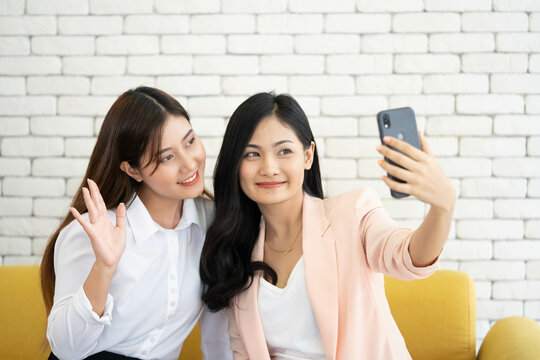 two happy ladies take selfie from smartphone on yellow couch and white brick wall background, lesbian girlfriend lovers enjoy and smile photo shooting by using cellphone, collage students show camera.