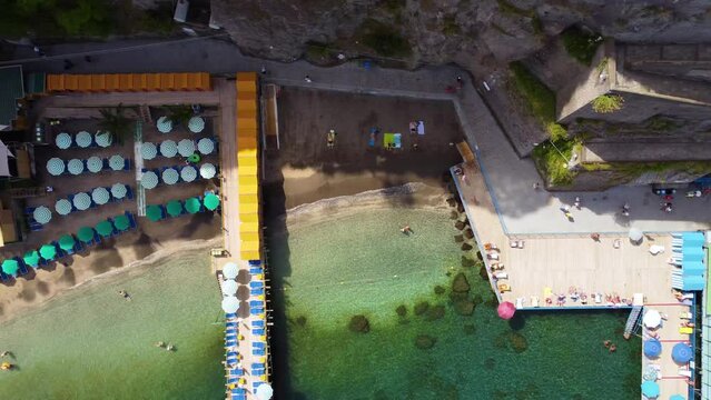 Aerial drone top down footage of Spiaggia Pubblica at steep cliff coastline of Sorrento, Campania, Italy. Summer beach resort platforms on with people sunbathing on sun lounger umbrellas from above