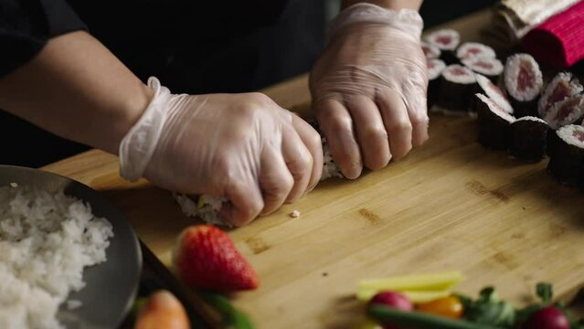Close-up shot of a japanese chef wearing gloves rolling sushi with ingredients inside it and shaping it with bamboo mat. Man cutting the roll with knife on wooden board
