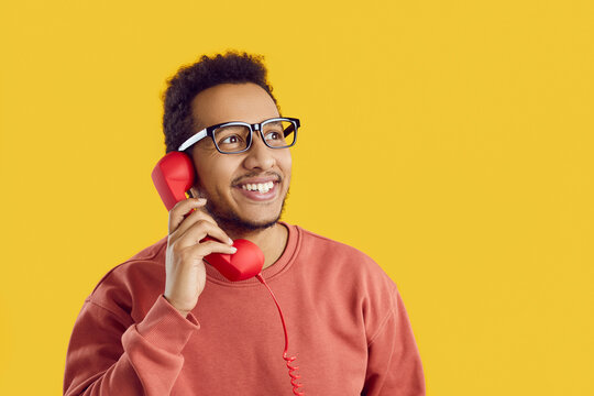 Cheerful man answers phone. Positive young black guy talking on landline telephone. Happy student standing on solid yellow background, holding red receiver, calling friends, looking away and smiling