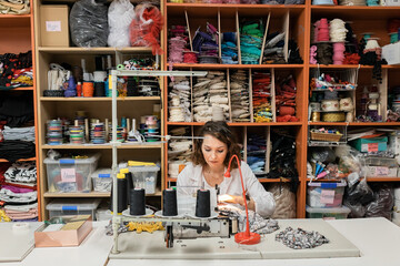 Concentrated seamstress woman sewing suit or fabrics at work table with sewing machine in workshop
