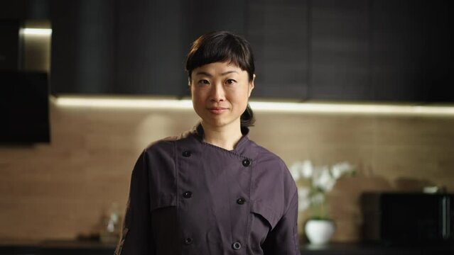 Medium shot of a smiling beautiful japanese female chef looking at camera while standing in home kitchen
