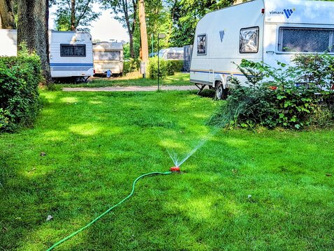Berlin, 2022: Caravans at a campsite in the forest on a green meadow with a lawn sprinkler in operation