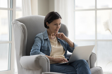 Focused mature freelance professional woman using online app on laptop for job communication,...