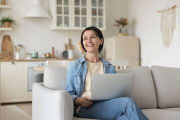 Happy mature freelancer woman holding laptop computer on lap, sitting on soft couch, looking away with positive thoughts, smiling, enjoying online communication, thinking, dreaming