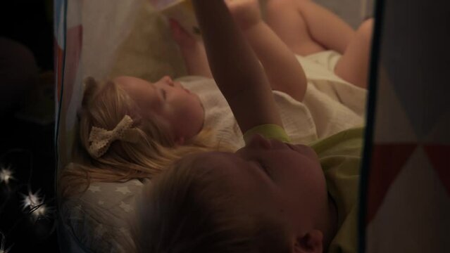 Little brother and sister 2 and 5 years old are lying on their backs in a children's tent at home and reading books. Happy little brother and sister are playing at home in a tent. High quality 4k