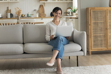 Obraz premium Happy attractive mature freelancer woman working at laptop at home, holding computer on lap, sitting on couch, talking on video call, looking at screen, laughing, enjoying Internet communication