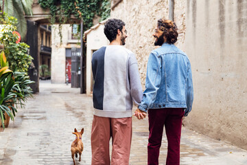 rear view of a gay male couple smiling happy walking with their little dog along a beautiful old street with plants, concept of lifestyle and love between people of the same sex - Powered by Adobe