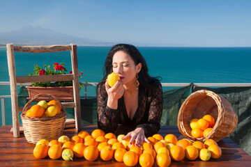 Mediterranean scenery: view of a typical mediterranean woman smelling oranges and lemons in a panoramic balcony in Sicily, with blue sea and Mount Etna in the background