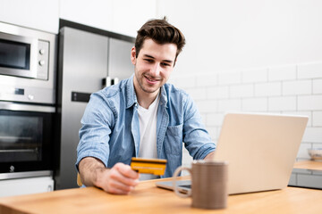 Man sitting in front of the computer holding a credit card at home - Young adult doing a online payment with his laptop at kitchen - business, technology concept