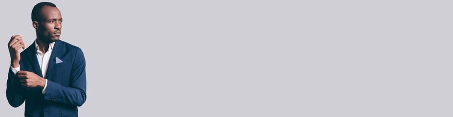 Elegant young African man in suit adjusting his sleeve while standing against grey background