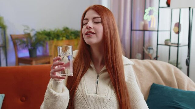 Thirsty redhead woman holding glass of natural aqua make sips drinking still water preventing dehydration sits at home living room. Girl with good life habits, healthy slimming, weight loss concept