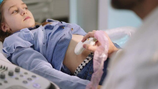 Hand of male doctor performing abdominal ultrasound on relaxed little girl who is lying on the bed in clinic