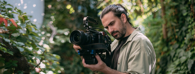A young male cameraman with a professional movie camera in his hands in a green garden.
