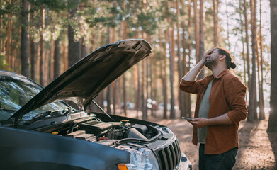 A frustrated man near a broken car with an open hood far outside the city in the woods.