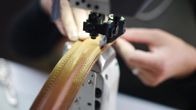 Woman making brown leather handbag and stitching handles on sewing machine at workplace in slow-motion. Close-up hands