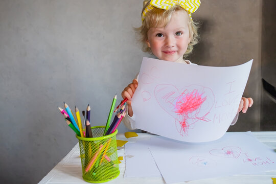 A little girl holds a piece of paper with a drawn congratulations for Mother's Day and an inscription I love mom
