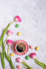 Pink porcelain cup with freshly brewed coffee on a light gray background, surrounded by tulips and colorful Easter eggs. Happy Easter and spring holiday concept. Top view. Place for text. Flat lay.