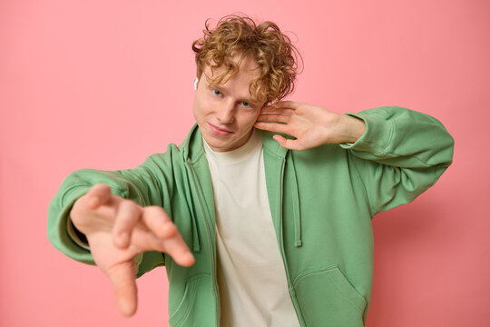 Horizontal shot of positive young man with curly red hair dances carefree waves hands moves with rhythm of music wears headphones dressed in green zipped hoodie isolated on pink background copy space.