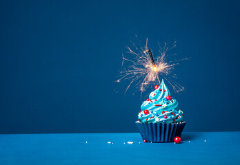 Blue cupcake with red and white sprinkles and lit sparkler on a blue background. - 580157760