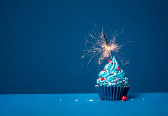 Blue cupcake with red and white sprinkles and lit sparkler on a blue background. © jfunk