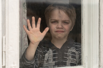 A dirty little boy is looking through an old dirty window. He put his hand to the window saying...
