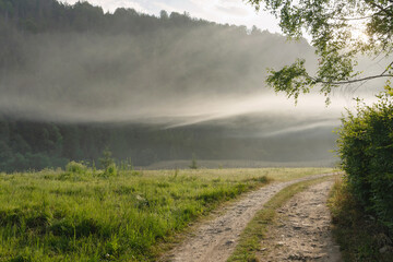 A dirt road in a mountain village leads to the unknown. Morning sunbeams and fog can be seen from around the bend (concept: the road to the unknown)