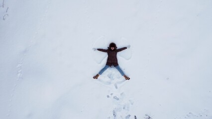 Naklejka premium A young woman lies on the snow and makes the figure of an angel. View of the girl from above. The camera moves away.