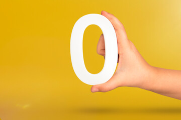 Numeral zero in hand. A hand holds a white number zero on a yellow background with copy space. Zero concept, 0 percent interest rate, minimum air emissions, cost or credit no increase.