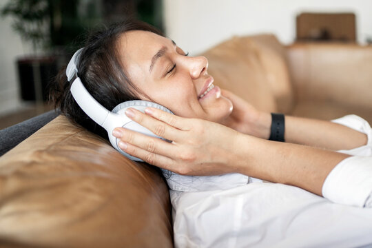 A woman listening to an audiobook with headphones is resting lying on the comfort house sofa.