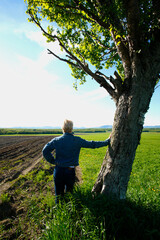 Rear view of mature woman leaning on tree outdoors overlooking freshly plowed, farmland in Spring.