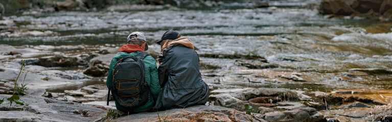 Rear of couple of hikers sitting on bank of rocky river and resting when hiking in mountains.