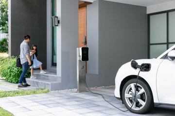 Focus progressive electric vehicle recharging at home charging station using clean and renewable energy with blurred father and daughter walking in background for future renewable energy concept. - obrazy, fototapety, plakaty