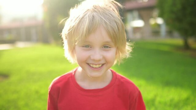 Slow motion close-up outdoors portrait of happy school-age child. Cute boy with blond hair emotionally laughs and looks at you. Real emotions of kids. Beautiful european child smiles and rejoices