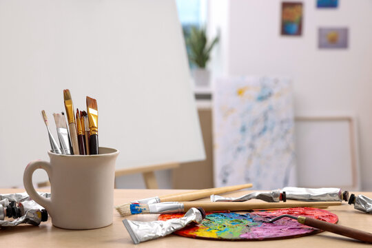 Art supplies on table near easel with canvas indoors. Space for text