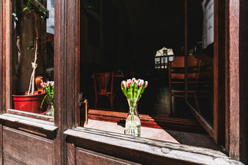 On a wooden window sill, a glass jar brims with crystal-clear water, cradling a bouquet of pink...