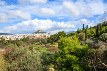 Fototapeta na wymiar Ruins can be seen among the trees at the ancient Greek Agora at the base of the Acropolis Hill with Mount Lycabettus in view in the distance, in Athens, Greece.