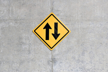 up and down arrows on yellow square diamond  traffic sign hanging on gray raw concrete wall, arrow indicates path that car can run in opposite direction, two way traffic sign