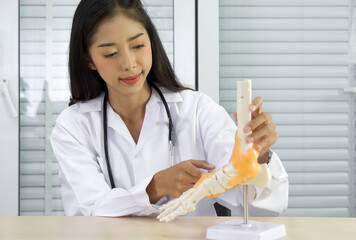 Young asian doctor in white gown and stethoscope pointing at human skeleton foot ankle bone joint...