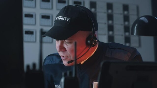 Security officer in headphones sits by table, looks view from security cameras displayed on monitor and talks with monitoring operator. Tracking system. Observation and CCTV technology. Portrait view.