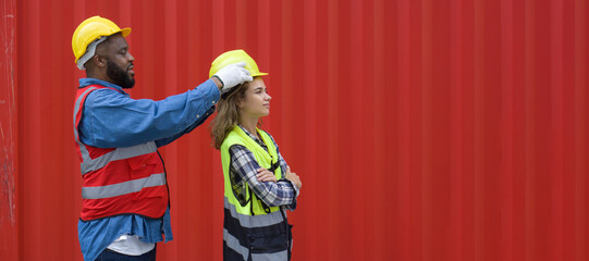 A foreman with mustache and beard helps a female apprentice wear a hardhat. Both of them wearing...