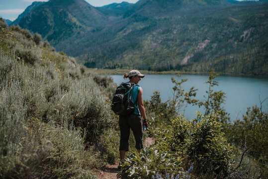 Rear view of female hiker with backpack standing on field amidst plants at lakeshore against mountains