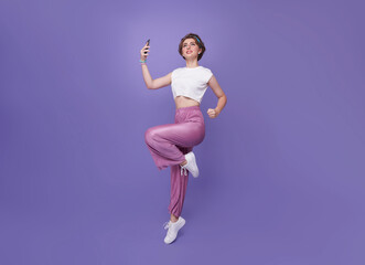 Happy young teen woman celebrating with mobile phone while jumping up on studio background. full body