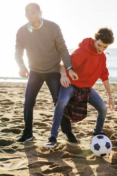 Full length of father and son playing soccer at beach during sunny day