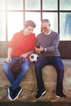 Happy father showing smart phone to son while sitting on retaining wall
