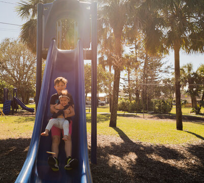 Brother with sister sliding on slide at playground during sunny day