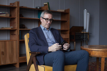 Confident elderly businessman with grey hair in suit and glasses working and typing on mobile phone indoors on background of office wall with boockshelf