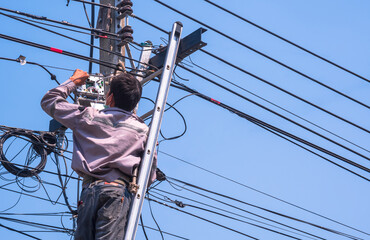 Technician on ladder is checking and renovating the CATV system hanging on black optical fiber cables for amplify TV signals on electric pole against blue sky background