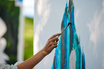 Artist hand holds paint brush and draws abstract surreal image on white canvas at outdoor art painting festival, paintings art picture process. Artist paints atmospheric picture - obrazy, fototapety, plakaty